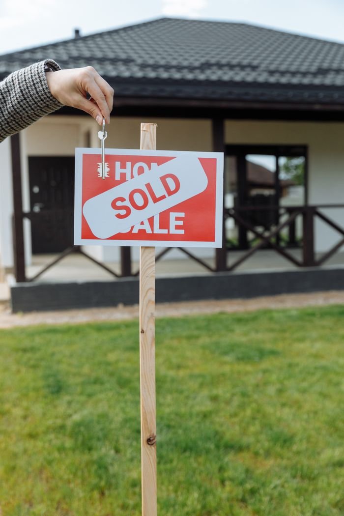 gallery-02 A hand holding a key in front of a sold sign and a house, symbolizing a successful real estate transaction.